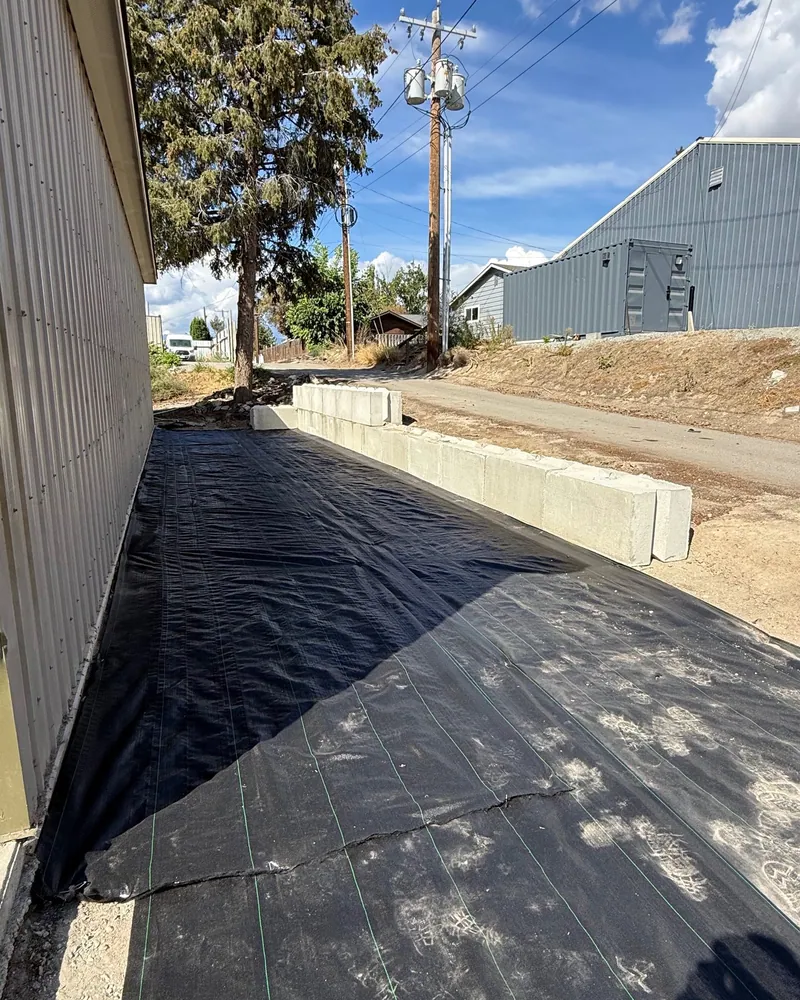 Black geotextile laid the length of the alley beside the metal building with concrete blocks along the road edge